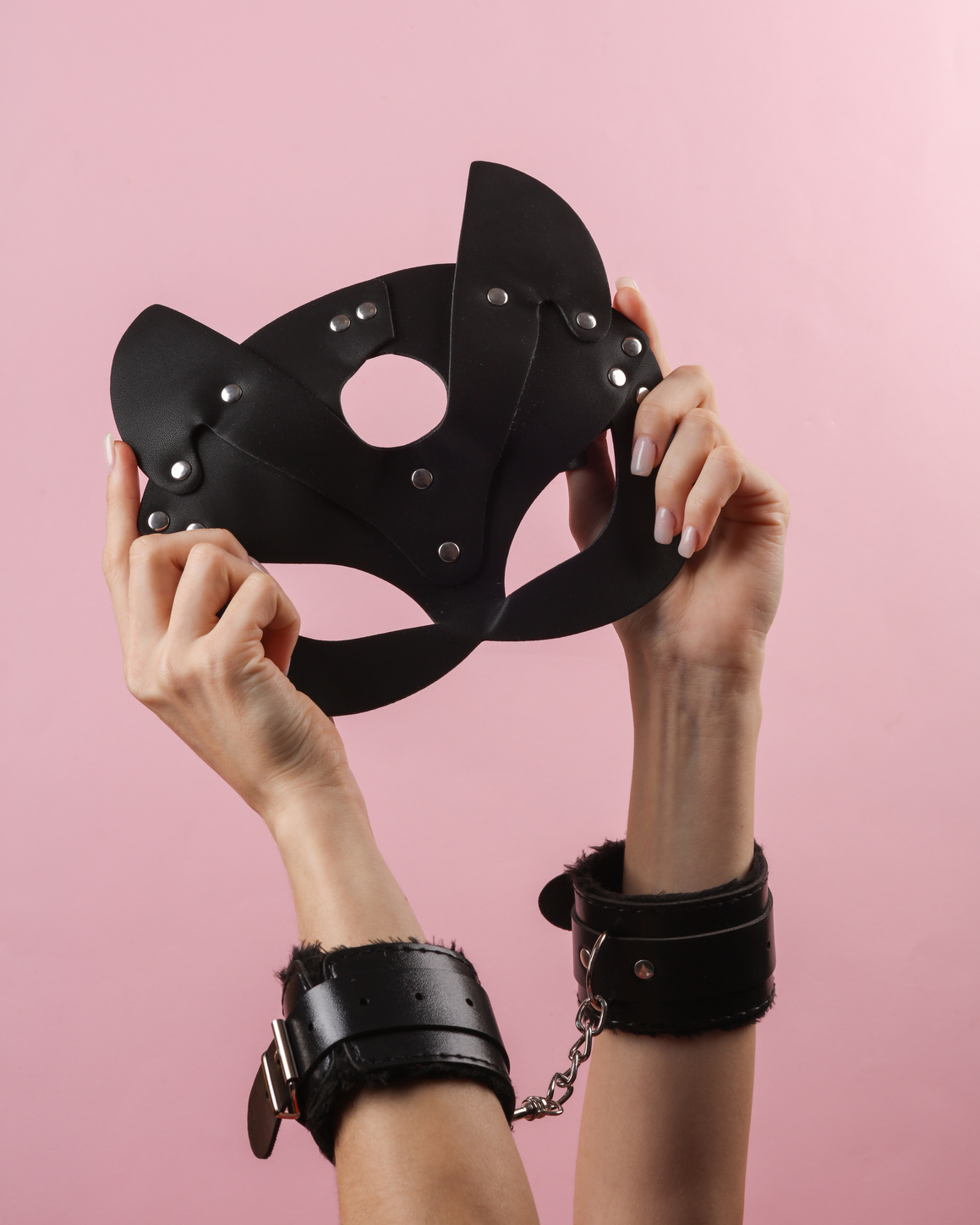 Women's hands in leather handcuffs holding black cat mask on pink background. BDSM accessories, domination, sex games
